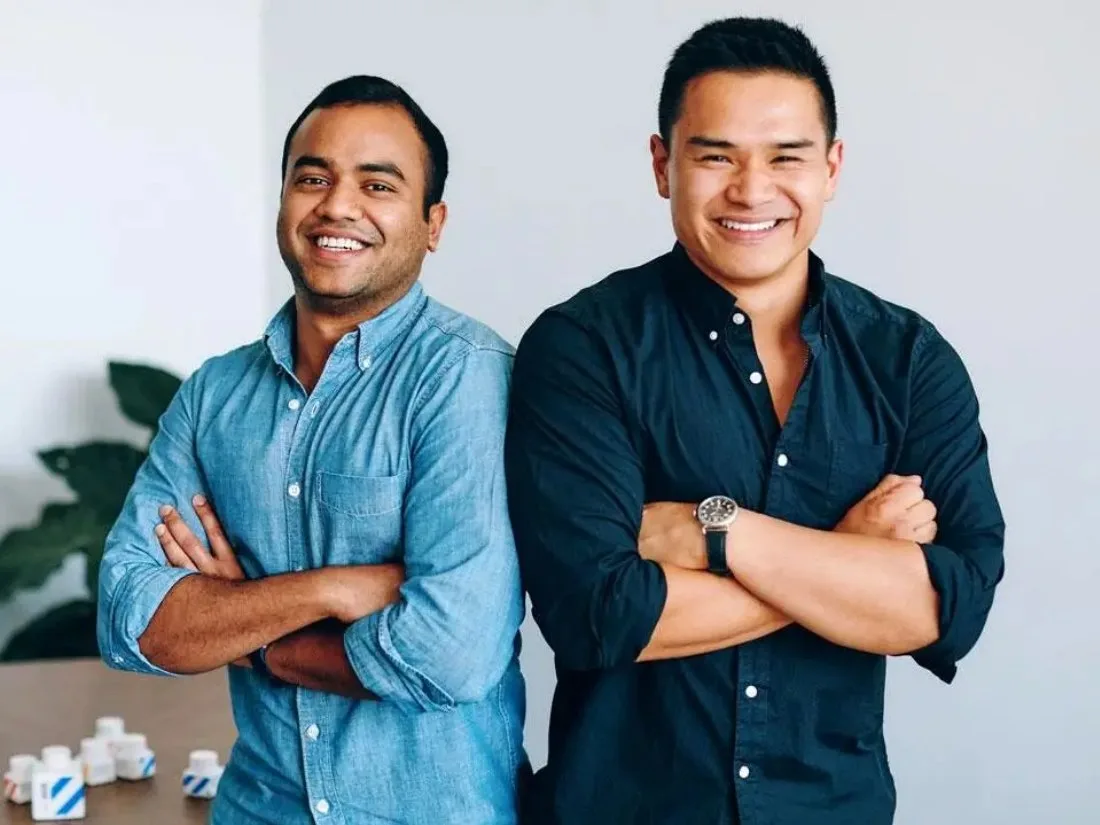 The image features two men standing side-by-side with arms crossed, smiling at the camera. There are small white bottles with blue stripes visible on a table in the lower-left corner.