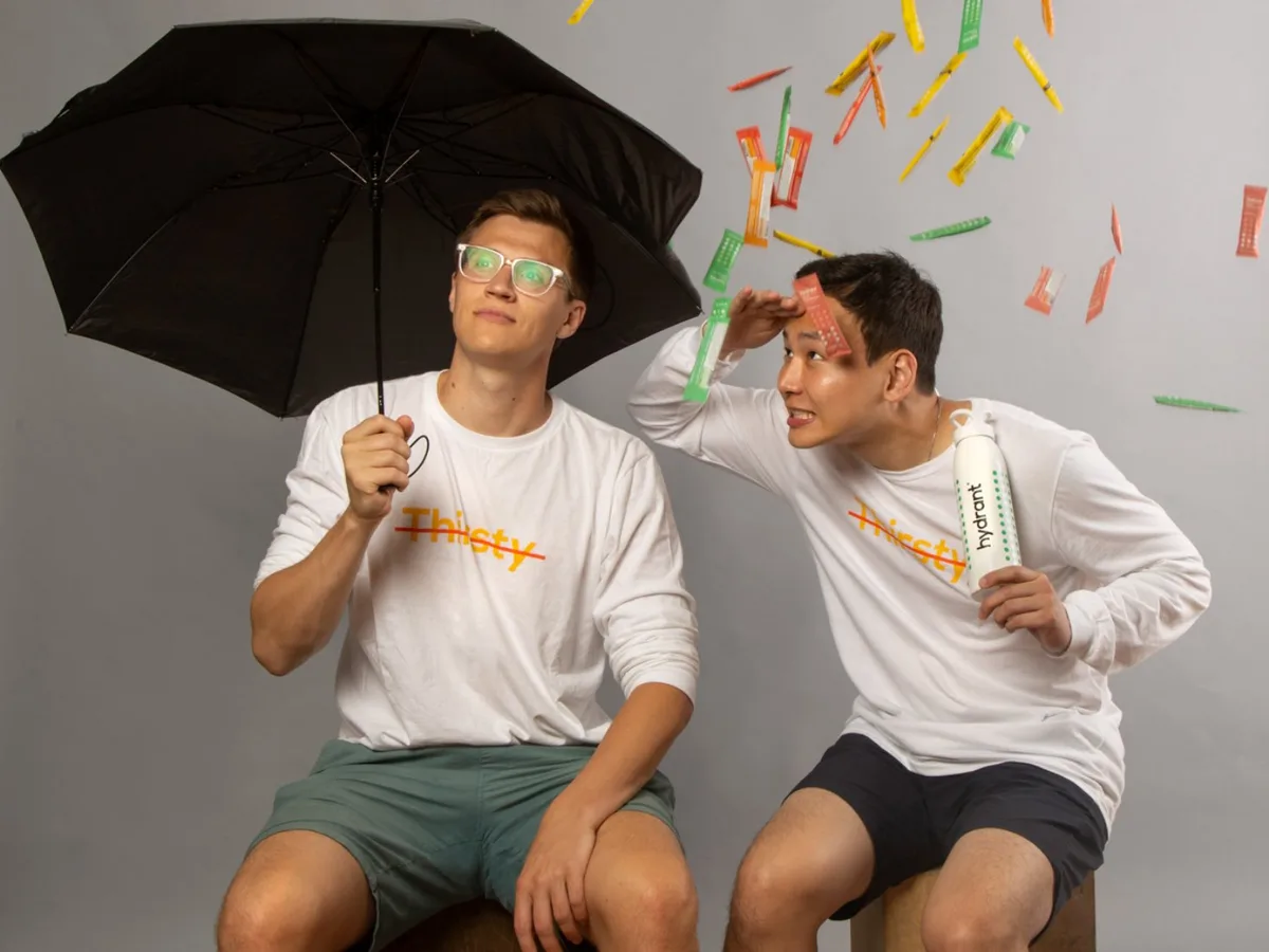 The image uses a visual pun where the word 'Thirsty' is crossed out on their shirts, and they are interacting with product packets falling like rain.